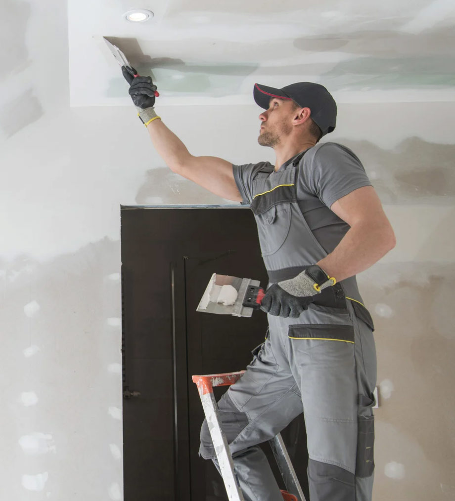 A worker on a ladder plastering a ceiling with a trowel in a partially finished room.