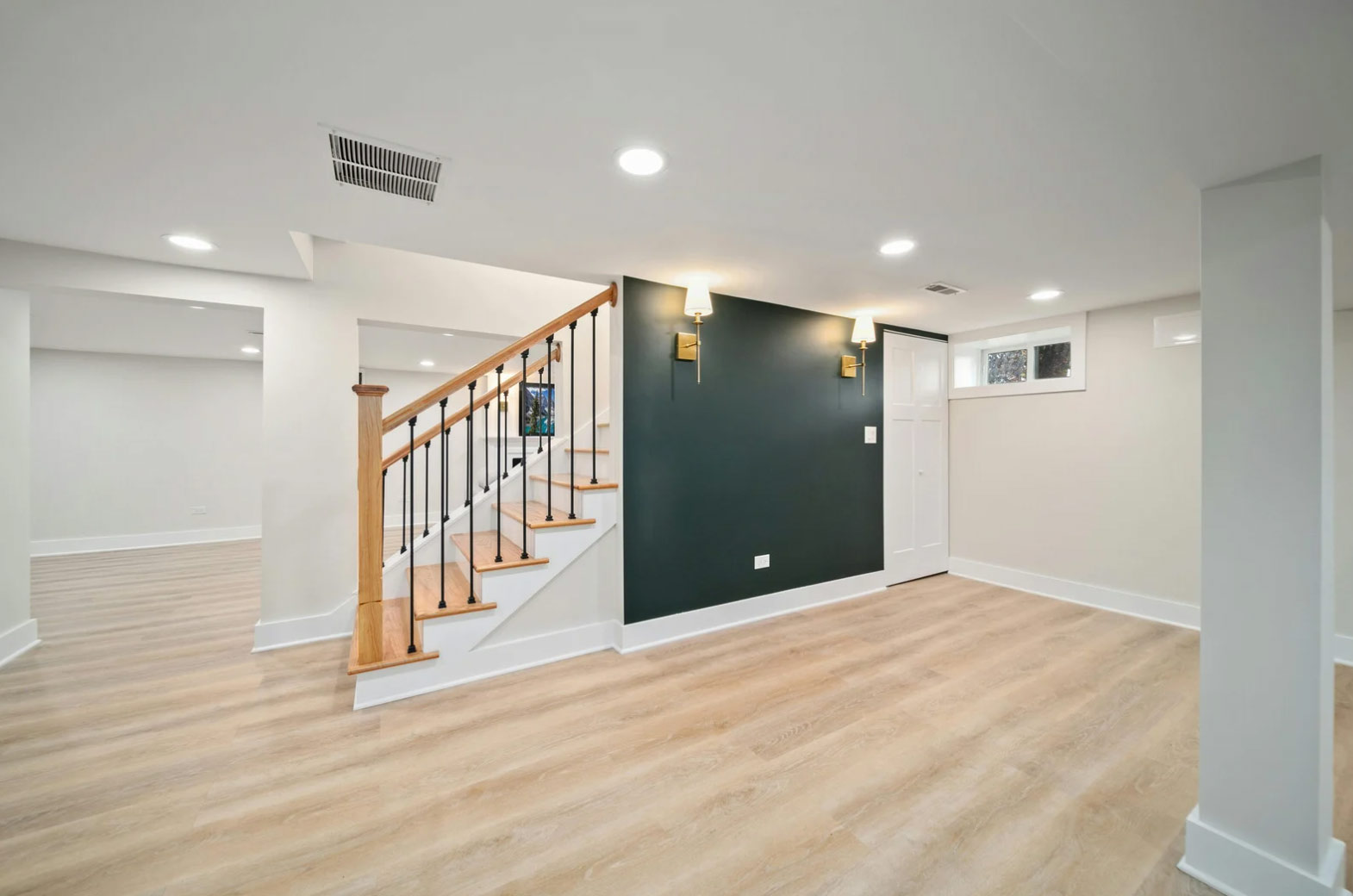 Modern finished basement with light wood floors, white walls, a dark accent wall, and a wooden staircase.