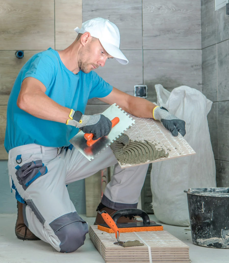 A worker applies adhesive to a tile with a notched trowel during a tiling project.