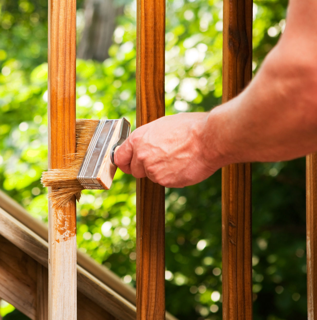 A person staining wooden railing with a paintbrush outdoors, surrounded by greenery.
