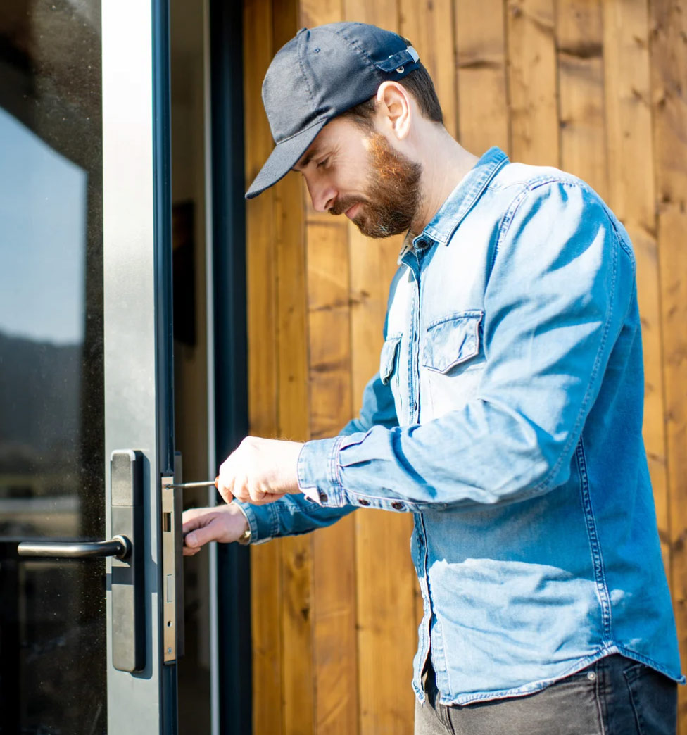 Man in a blue denim shirt and cap unlocking a glass door with a screwdriver.