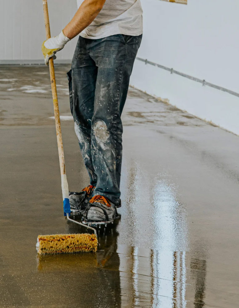 Person applying epoxy coating to a concrete floor with a roller in an indoor space.