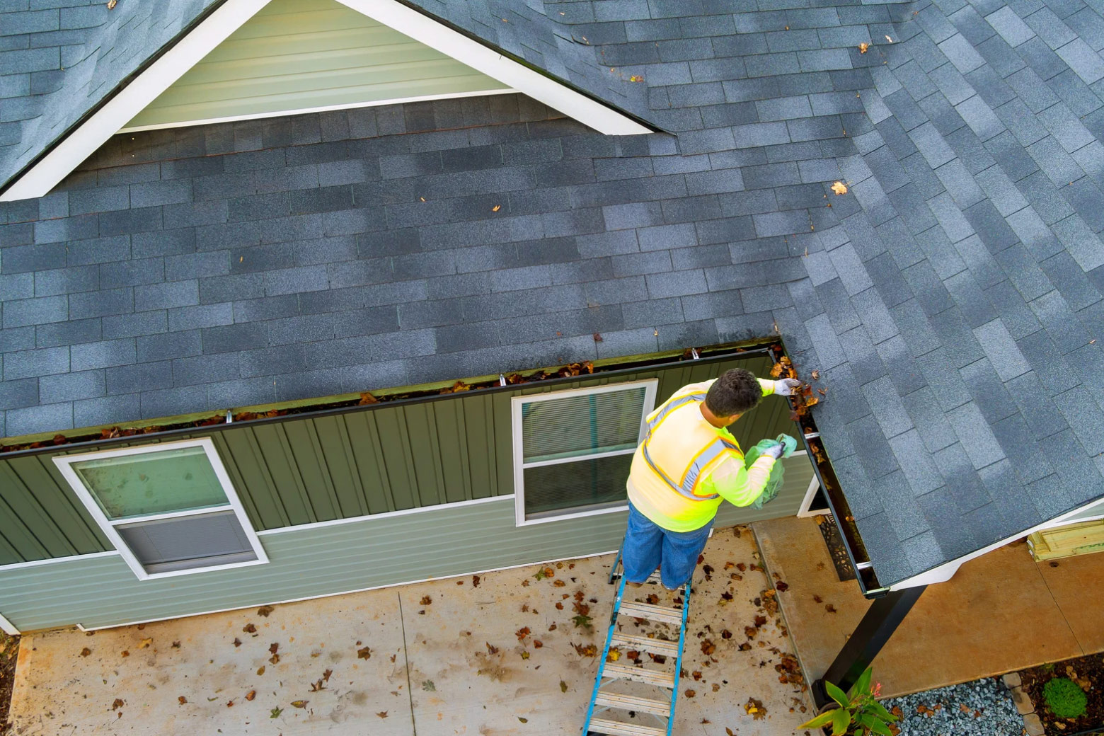 A worker on a ladder cleans leaves from a house gutter while wearing a yellow safety vest.