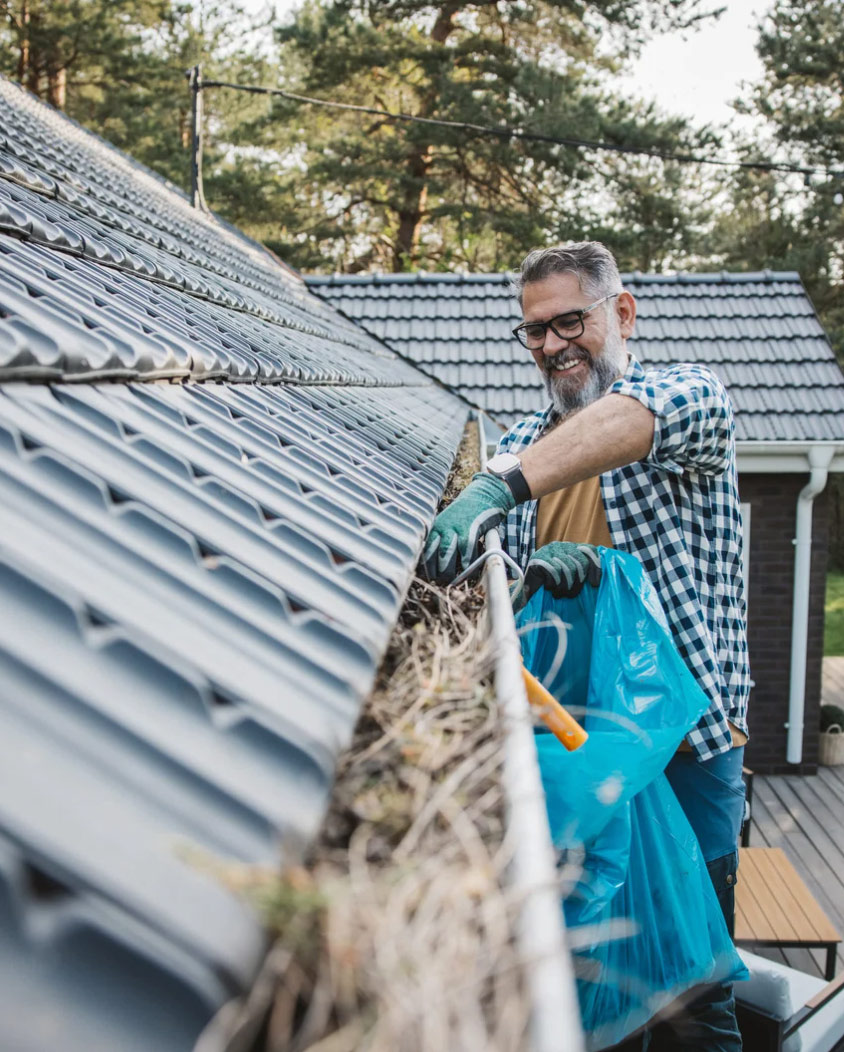 Smiling man in gloves cleaning leaves from a house gutter into a blue bag on a rooftop.