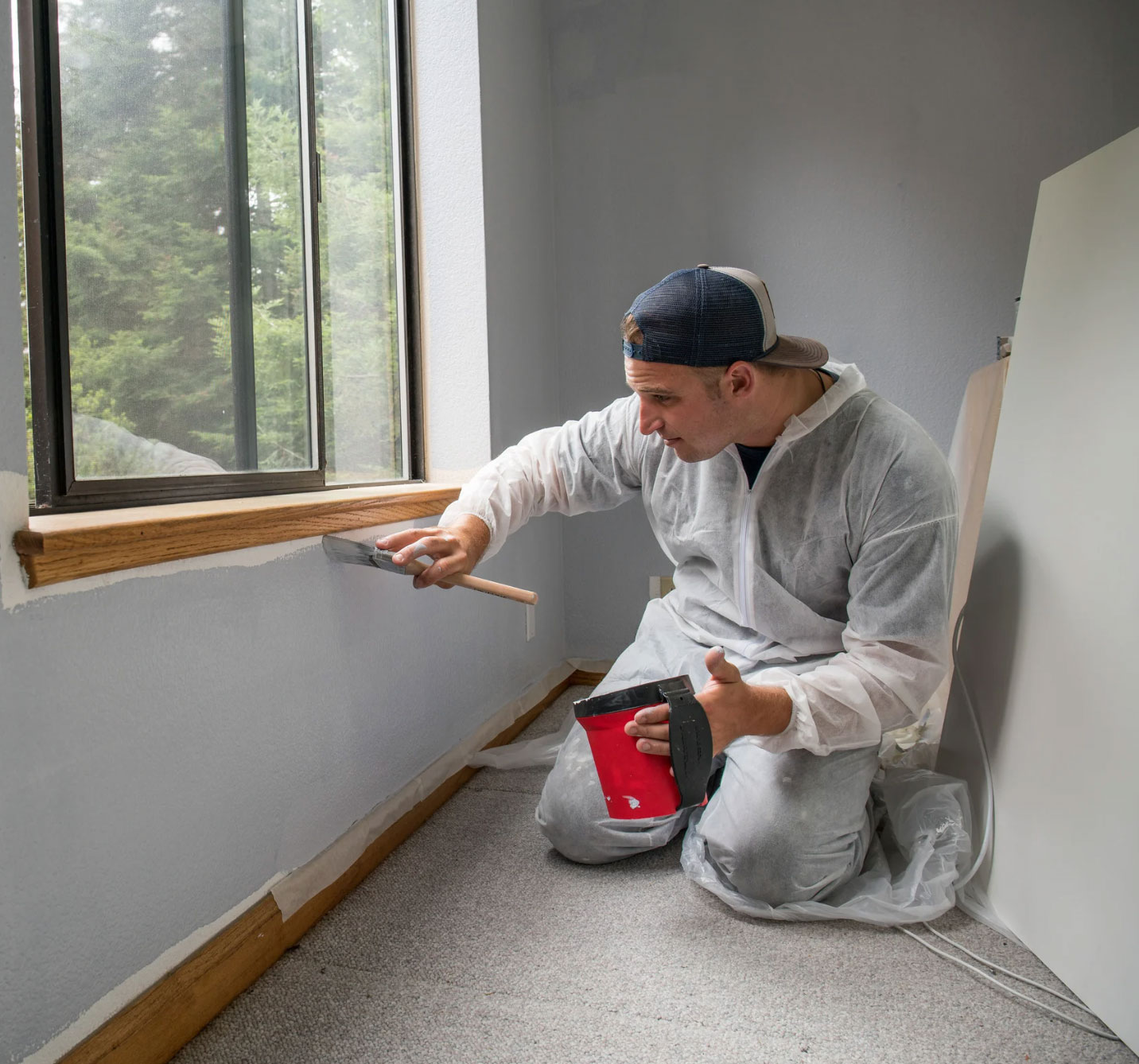 Man in coveralls spackling a wall near a window with trees visible outside, holding a putty knife and tool.