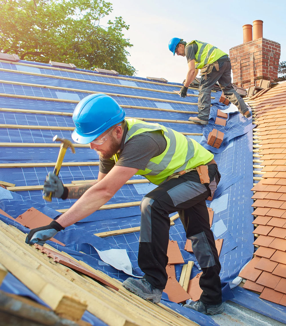 Two construction workers in safety gear installing tiles on a sloped roof.