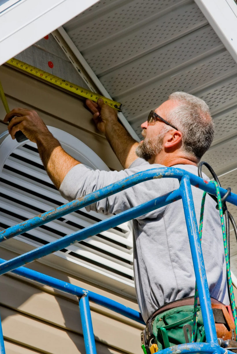 A man on a lift measures siding on a house exterior with a tape measure, wearing sunglasses and tools.