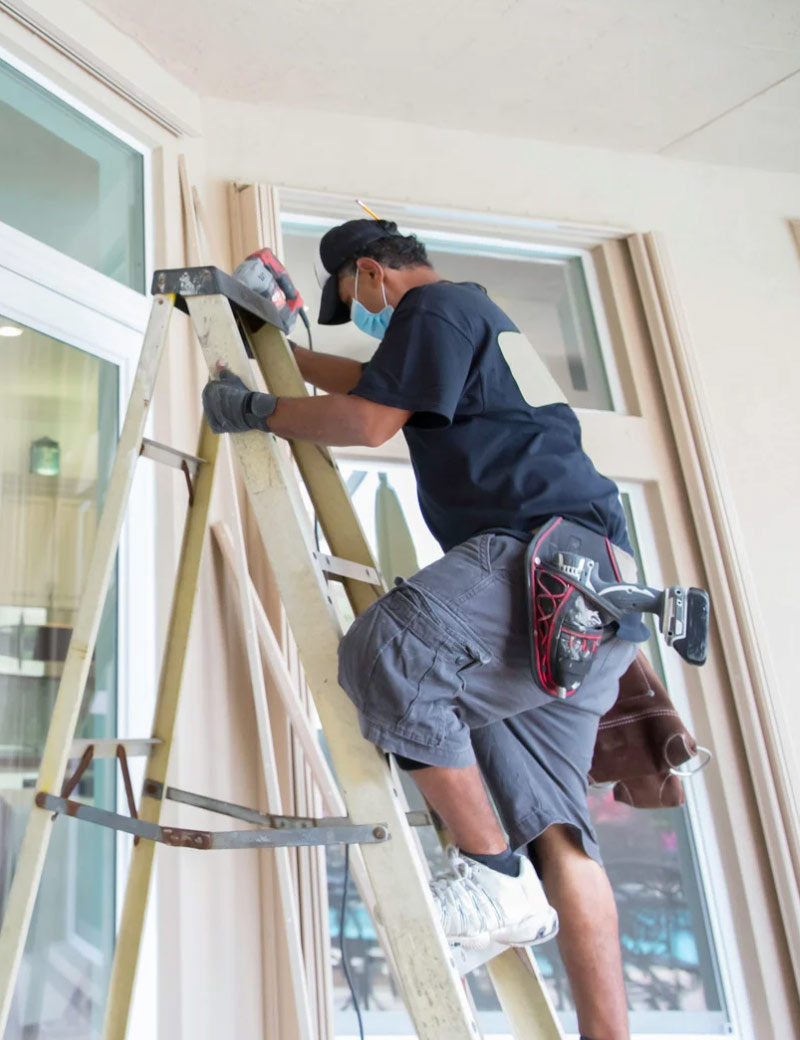 A man on a ladder installs window frames, wearing a mask, gloves, and tool belt.